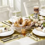 تصویری از یک میز ناهارخوری شیک گل آرایی شده با ست کامل ظرف و قاشق و چنگال با ترکیب رنگ سفید و طلایی که ما رو یاد بینوایان ژان وار ژان میندازه و مربوط به موضوع میز غذاخوری خانگی شیک هستش.